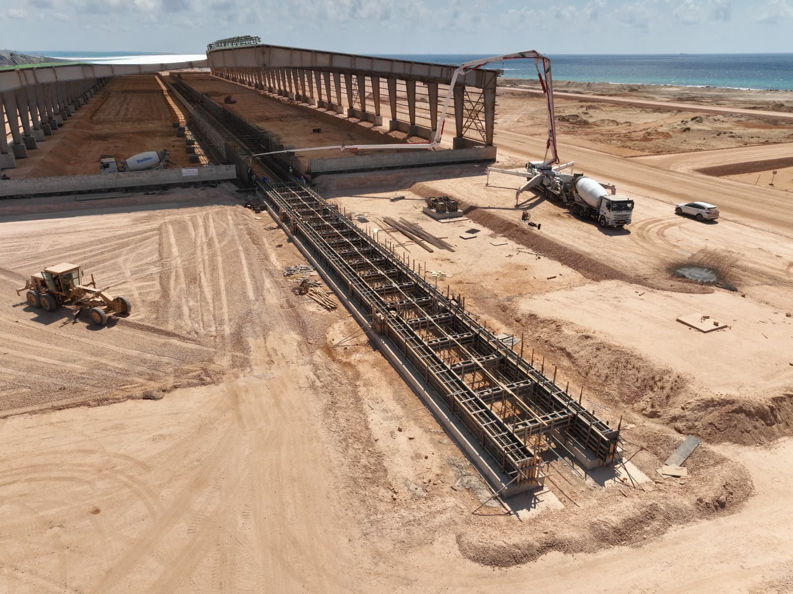 Aerial view of an industrial construction site near the sea with concrete structures, a concrete pump truck, a mixer, and earthmoving equipment.